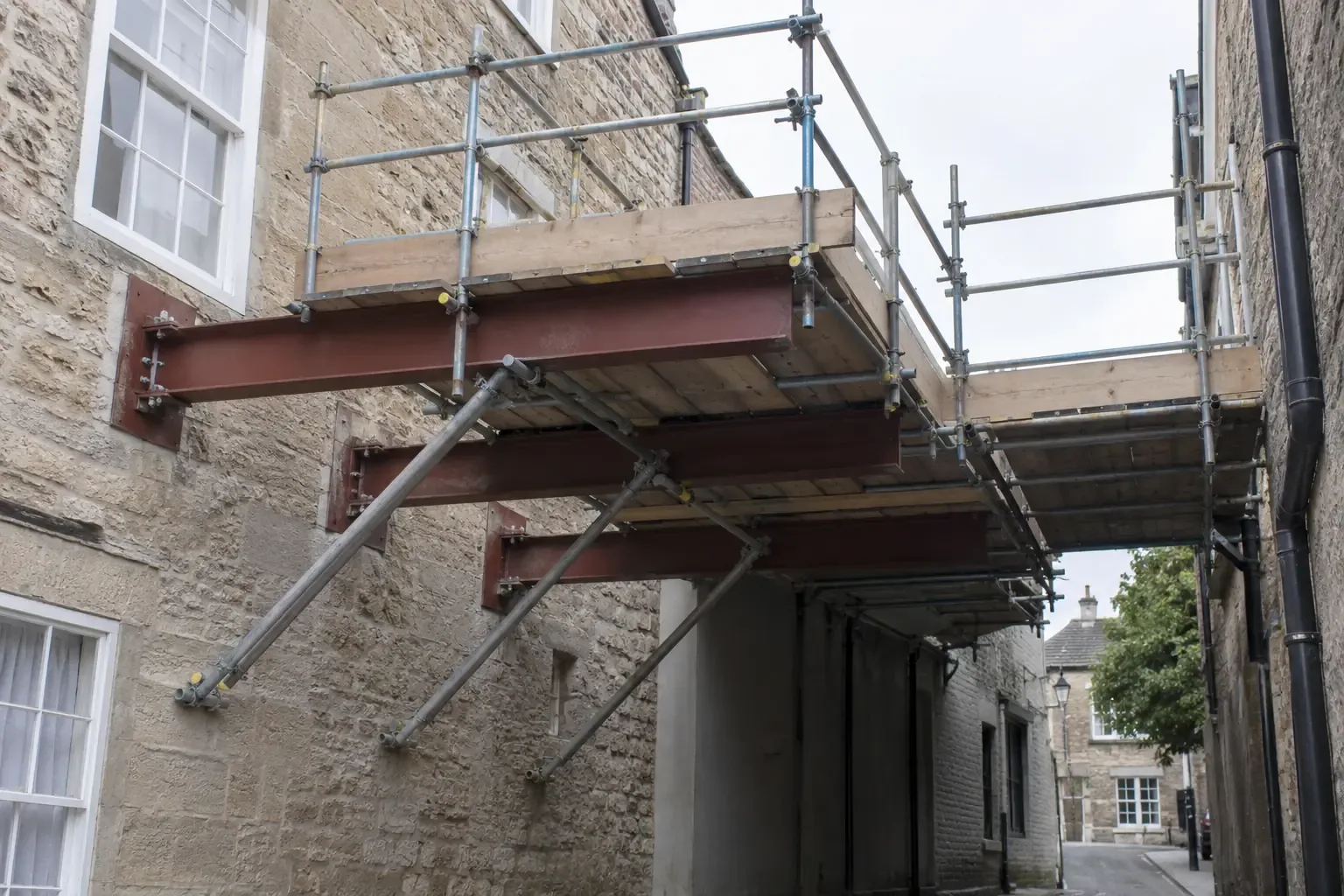 Cantilever scaffolding — A clearly engineered cantilever scaffold working deck projecting from substantial anchored horizontal steel needle beams through a masonry building facade above a narrow covered passage where ground supports cannot stand; triangulated braces, full guardrails and toe boards, no workers.