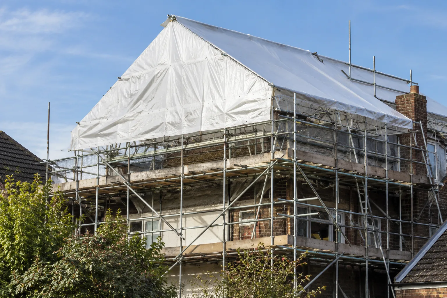 Temporary roof scaffold protecting a British residential renovation, white weatherproof sheeting stretched over a pitched temporary scaffold roof, robust metal tube support and complete edge-protected platforms, realistic properly braced construction.